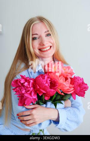 Lächelnde junge Frau mit hellrosa Pfingstrosen in minimalistischem Studio-Porträt mit weichem natürlichem Licht und frischer Frühlingsstimmung. Stockfoto