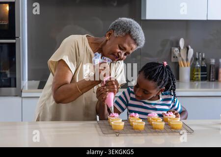Afrikanische Familie Senior Frau führt rosa Paspelbeutel und Jugendmädchen hält Cupcake in der Küche Stockfoto