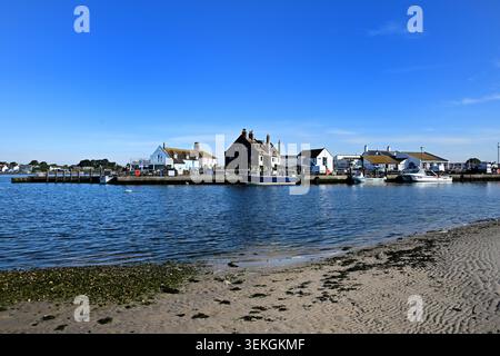 Mudeford Quay, Christchurch Harbour in Dorset, England, von der Sandküste aus gesehen an einem ruhigen, sonnigen Tag mit Fischerbooten und Hafengebäuden Stockfoto