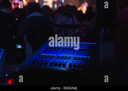 Blau beleuchtetes Soundboard und Beleuchtungssteuerung bei einem Live-Konzert, Techniker mischen Audio und Licht aus dem hinteren Teil eines dunklen Veranstaltungsortes, während das Publikum zusieht Stockfoto