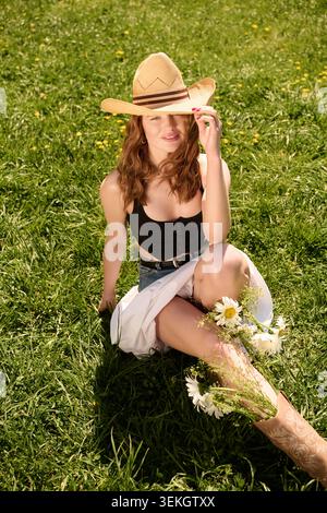 Eine junge Frau in Sommerkleidung sitzt auf einem grünen Feld und lächelt mit einem Strohhut. Stockfoto