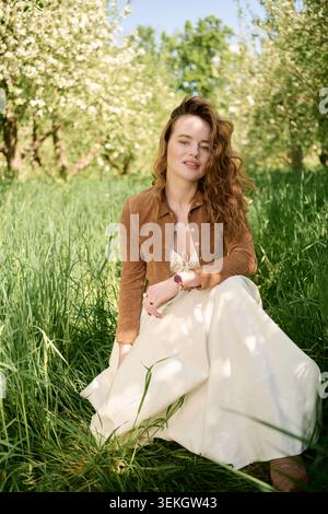 Eine anmutige junge Frau sitzt in einem üppigen grünen Feld, umgeben von blühenden Bäumen. Stockfoto