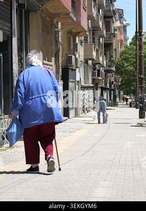Eine ältere Frau, die mit einem Stock weggeht und eine Tasche auf einer Stadtstraße trägt, repräsentiert Unabhängigkeit, Alltag und das Vergehen der Zeit in einer Stadt Stockfoto