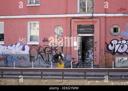 Lieferer in Schutzhelm und Uniform mit seinem Fahrrad, der eine gelbe Thermotasche vor einem Gebäude mit abblätternder Farbe, Graffiti, A sichert Stockfoto