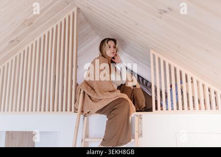 Nachdenkliche Junge Weiße Frau, Die In Einer Hölzernen Loft-Hütte Sitzt Stockfoto