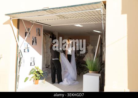 Zwei Frauen in einem Modestudio passen ein Hochzeitskleid an eine Schaufensterpuppe an. Nähmaterialien und Kleidermuster sind im Hintergrund zu sehen Stockfoto