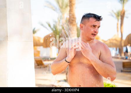 Ein kaukasischer Mann steht unter einer Stranddusche im Freien und Wasser fließt über seinen Körper in der Nähe von Palmen und Resorts. Stranderholung Hygiene va Stockfoto