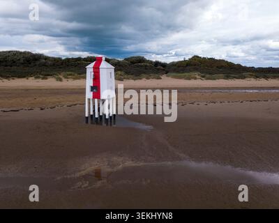Drohnenblick auf den Burnham‑on‑Sea Low Lighthouse am Sandstrand, der seine markante rot-weiße Struktur vor Dünen und Himmel einfängt Stockfoto