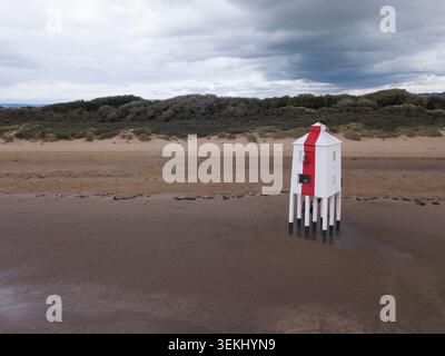 Drohnenblick auf den Burnham‑on‑Sea Low Lighthouse am Sandstrand, der seine markante rot-weiße Struktur vor Dünen und Himmel einfängt Stockfoto
