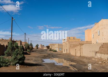 Straßenszene in Merzouga, Marokko, mit Schlammgebäuden, Versorgungsmasten und einer Pfütze auf der unbefestigten Straße unter klarem blauem Himmel. Stockfoto