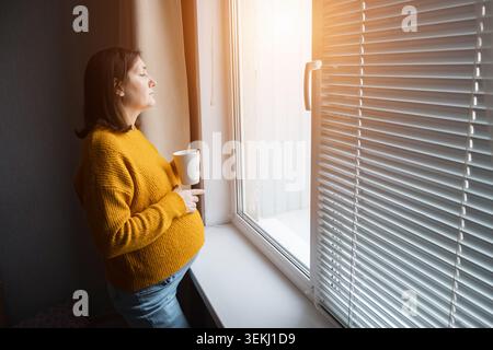 Schwangere Frau in gelbem Pullover, die eine Tasse an einem Fenster mit warmem Morgenlicht hält Stockfoto