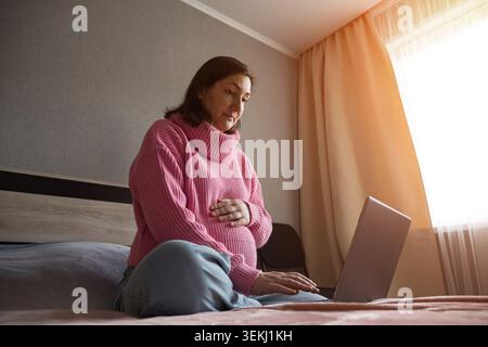 Schwangere Frau sitzt zu Hause auf dem Bett mit einem Laptop und hält ihren Bauch Stockfoto