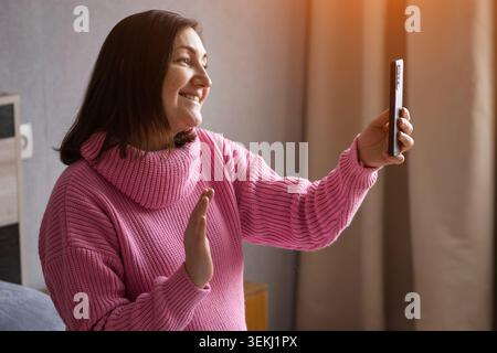 Lächelnde schwangere Frau, die ein Smartphone in einem gemütlichen Wohnzimmer hält Stockfoto