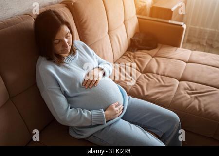 Schwangere Frau ruht auf einem beigen Sofa in einem warmen Wohnzimmer Stockfoto