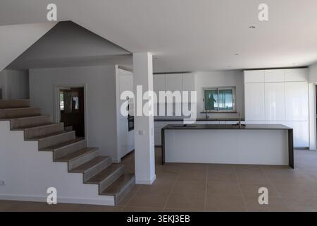 Ein zeitgenössisches Innenleben mit einer minimalistischen weißen Küche, einer großen Insel und einer markanten Treppe, die zu einer anderen Ebene führt Stockfoto