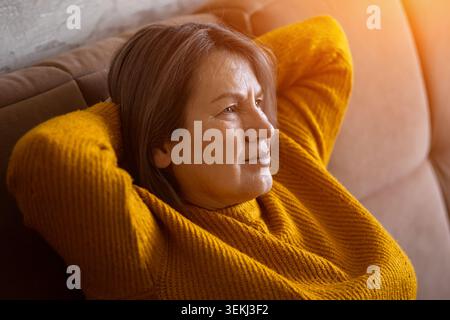 Nachdenkliche ältere Frau, die sich zu Hause im warmen Nachmittagslicht auf einem Sofa entspannt Stockfoto