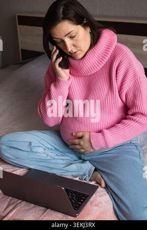 Schwangere Frau, die in einem rosa Pullover auf dem Bett sitzt, telefoniert, während sie ihren Bauch hält Stockfoto