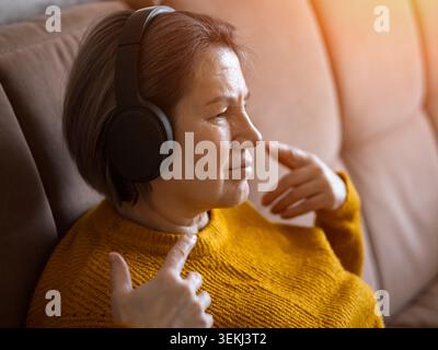 Ältere Frau mit Kopfhörern, die sich bei warmem Licht auf einem Sofa entspannen Stockfoto