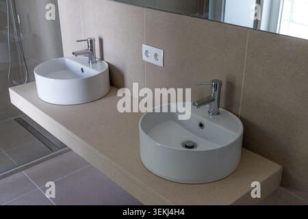 Das moderne Badezimmer verfügt über zwei elegante, weiße Waschbecken, die auf einer schwebenden Arbeitsplatte mit Chromarmaturen und einer gefliesten Wand montiert sind Stockfoto