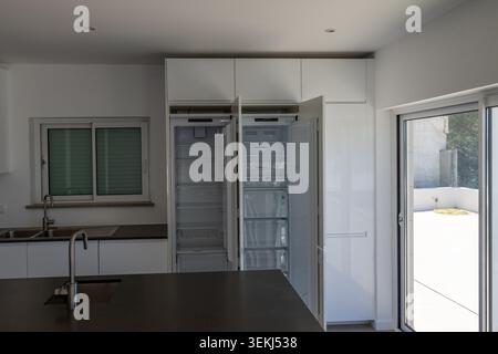 Ein moderner Küchenbereich mit offenem Kühlschrank, integrierten weißen Schränken und einer dunklen Arbeitsfläche Stockfoto