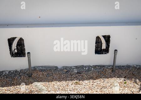 Unbearbeitetes Gebäude außen mit Kabelkanälen und Kabeln, die aus Wandöffnungen herausragen, wobei Rohre aus dem Boden darunter ragen Stockfoto