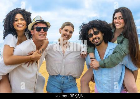 Vielfältige Gruppe von jungen erwachsenen Freunden lächelnd und genießt Sommerurlaub an einem Sandstrand unter blauem Himmel, mit zwei Paaren, die Huckepack haben, feiern Stockfoto