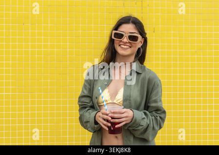 Eine junge Frau lächelt, trägt eine Sonnenbrille und ein lässiges Hemd über einem Badeanzug und hält ein erfrischendes Getränk mit einem Strohhalm vor einem leuchtend gelben Fliesenwal Stockfoto