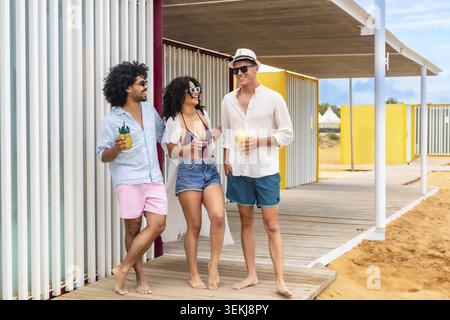 Eine Gruppe glücklicher junger Freunde lacht und hat Spaß, während sie bunte tropische Getränke halten und barfuß auf einer hölzernen Promenade am Strand stehen Stockfoto