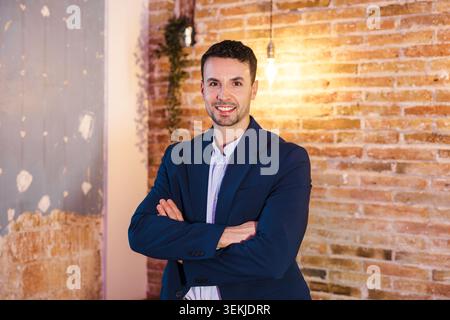 Selbstbewusster kaukasischer Unternehmer, der mit gekreuzten Armen im modernen Büro lächelt Stockfoto