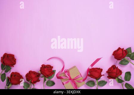 Eine wunderschön verpackte Geschenkbox mit einem rosafarbenen Band, umgeben von roten Rosen auf rosa Hintergrund, symbolisiert Liebe und Wertschätzung zum Muttertag Stockfoto