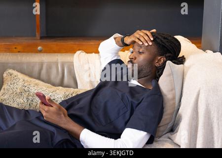 Afrikanischer Mann, der sich auf einem beigen Sofa in der Lounge liegend, mit marineblauen medizinischen Peelings, die ein rotes Smartphone halten. Couch, blau, Telefon, kontemplativ, Ermüdung, Innenraum Stockfoto