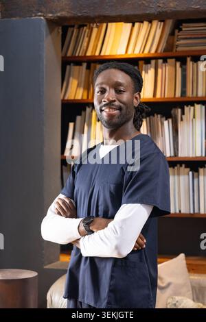Erwachsener afrikanischer Mann, der mit überkreuzten Armen im Arbeitszimmer steht, in marineblauen Schutteln und in Bücherregalen beobachtet. Arzt, medizinisch, Bibliothek, warm, ungekühlt, Holz, Stockfoto