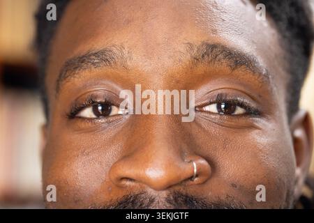 Afroamerikaner, der in die Kamera blickt, zeigt silberne Creppnasenpiercings. Nahaufnahme, Porträt, Skintexture, Gesichtsbehaarung, Stoppeln, Augenlider Stockfoto