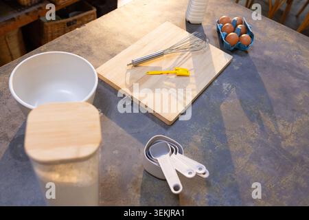 Das hölzerne Schneidebrett hält den Schneebesen und den gelben Schaber auf der abgenutzten Arbeitsplatte neben dem Mehlbecher und den Eiern Stockfoto