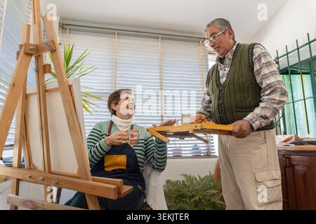 Ein fürsorglicher älterer Mann bringt Kaffee und Frühstück zu seiner Frau, während sie in einem hellen Kunststudio malt. Stockfoto