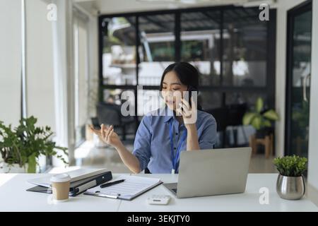 Junge Geschäftsfrau asiatisch, die mit dem Kunden über das Mobiltelefon auf dem Schreibtisch im Büro spricht Stockfoto