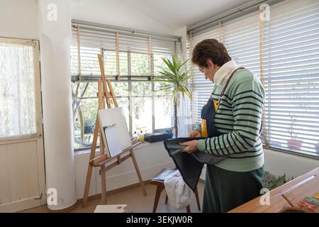 Eine ältere Frau in gestreiftem Pullover untersucht oder faltet eine dunkle Denim-Schürze, während sie in ihrem sonnendurchfluteten Studio neben einer Staffelei aus Holz und weißem Canvas steht. Stockfoto