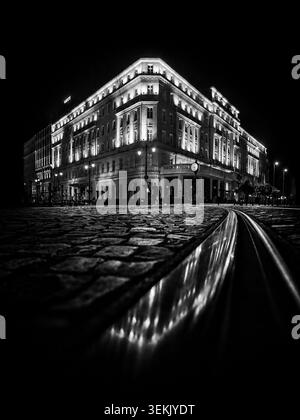Schwarz-weiße Nachtansicht auf das Carlton Hotel in Bratislava, Slowakei, mit leuchtender Fassade, Kopfsteinpflaster und Straßenbahnlinien. Stockfoto