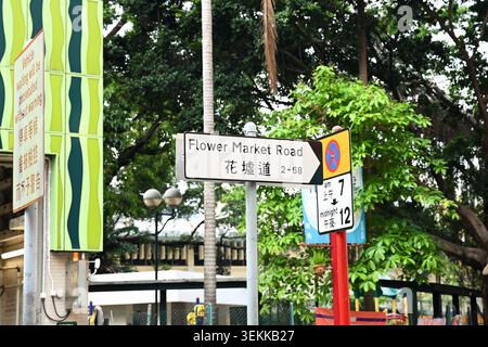 HONGKONG, CHINA, 21. April 2026, ein zweisprachiges Straßenschild für die Flower Market Road steht unter grünen Bäumen mit einer Warntafel auf der linken Seite. Stockfoto