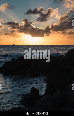 Felsiges Ufer mit Segelboot und spektakulärem Sonnenuntergang hinter dem Horizont Stockfoto