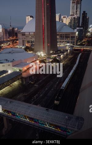 Blick aus der Vogelperspektive auf die Innenstadt von Toronto in der Abenddämmerung mit Union Station Zugkorridor, CN Tower, Rogers Centre, Pendlerzug und einer beleuchteten Fußgängerbrücke Stockfoto