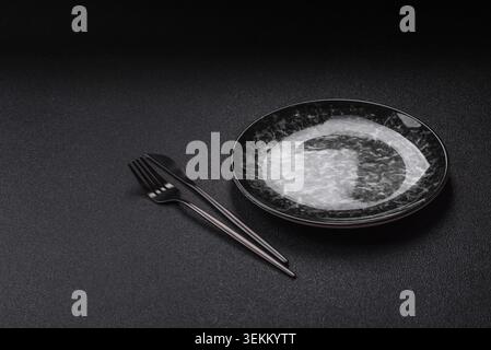 Leere runde schwarze Platte für Essen, Küchenutensilien auf Esstisch. Leere Platte auf dunklem Hintergrund Stockfoto