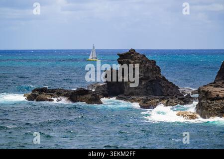 Zerklüftete Felsen erheben sich aus den Meeresgewässern mit einem Segelboot, das durch das Meer navigiert. Stockfoto