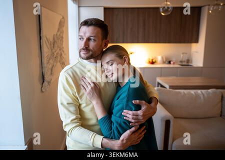 Positiv nachdenklicher Mann und Frau, die Fenster mit nachdenklichem Ausdruck umarmen und betrachten Stockfoto