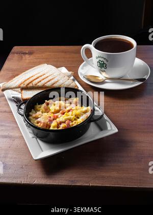 Gemütliches Frühstück auf dunklem Holztisch. Rührei mit Schinken in einer schwarzen gusseisernen Pfanne. Weißer Toast liegt neben der Schüssel, leicht getoastet. Becher Stockfoto
