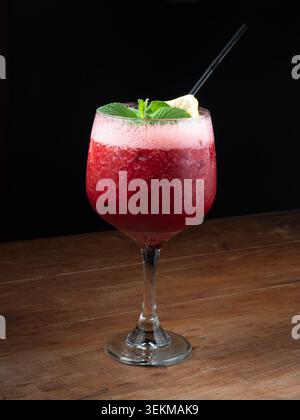 Roter Cocktail mit Minz- und Limettengarnitur auf rustikalem Holz. Schwarzer Strohhalm liegt auf der schaumigen Oberfläche. Dunkler Hintergrund sorgt für lebendige Getränkefarben. Stockfoto