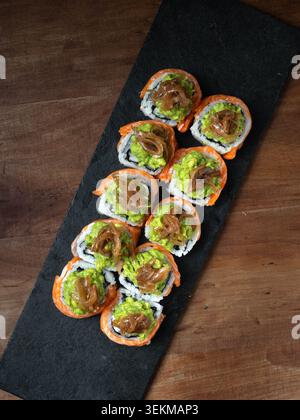 Sushi-Rollen liegen ordentlich auf einem eleganten schwarzen Schiefer. Jedes Brötchen verfügt über lebhafte Avocado- und herzhafte Beläge. Sanfte Beleuchtung hebt den frischen, glänzenden Textil hervor Stockfoto