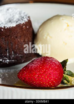 Reichhaltiger Schokoladenkuchen, mit Puderzucker bestäubt. Samtiges Vanilleeis neben dem Kuchen. Frische Erdbeere mit grünem Stiel liegt im Vordergrund. Servo Stockfoto