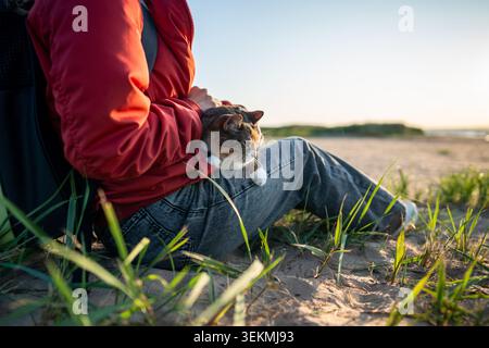 Der Reisende sitzt am Sandstrand und hält die Calico-Katze. Konzept der Reise mit Haustier, Freiheit, Outdoor-Lifestyle und Gesellschaft. Warmes Licht bei Sonnenuntergang, entspannt Stockfoto