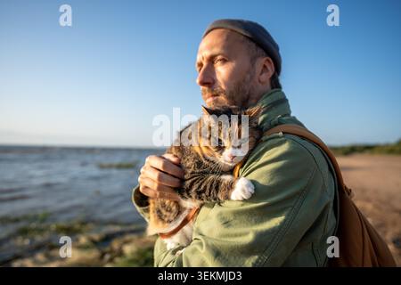 Ein fokussierter Mann hält die Katze in den Händen, während die Natur an der Küste spaziert. Haustierreisender. Katzenbesitzer umarmt flauschiges Kätzchen draußen. Tabby Calico Katze mit Leine sitzt Stockfoto